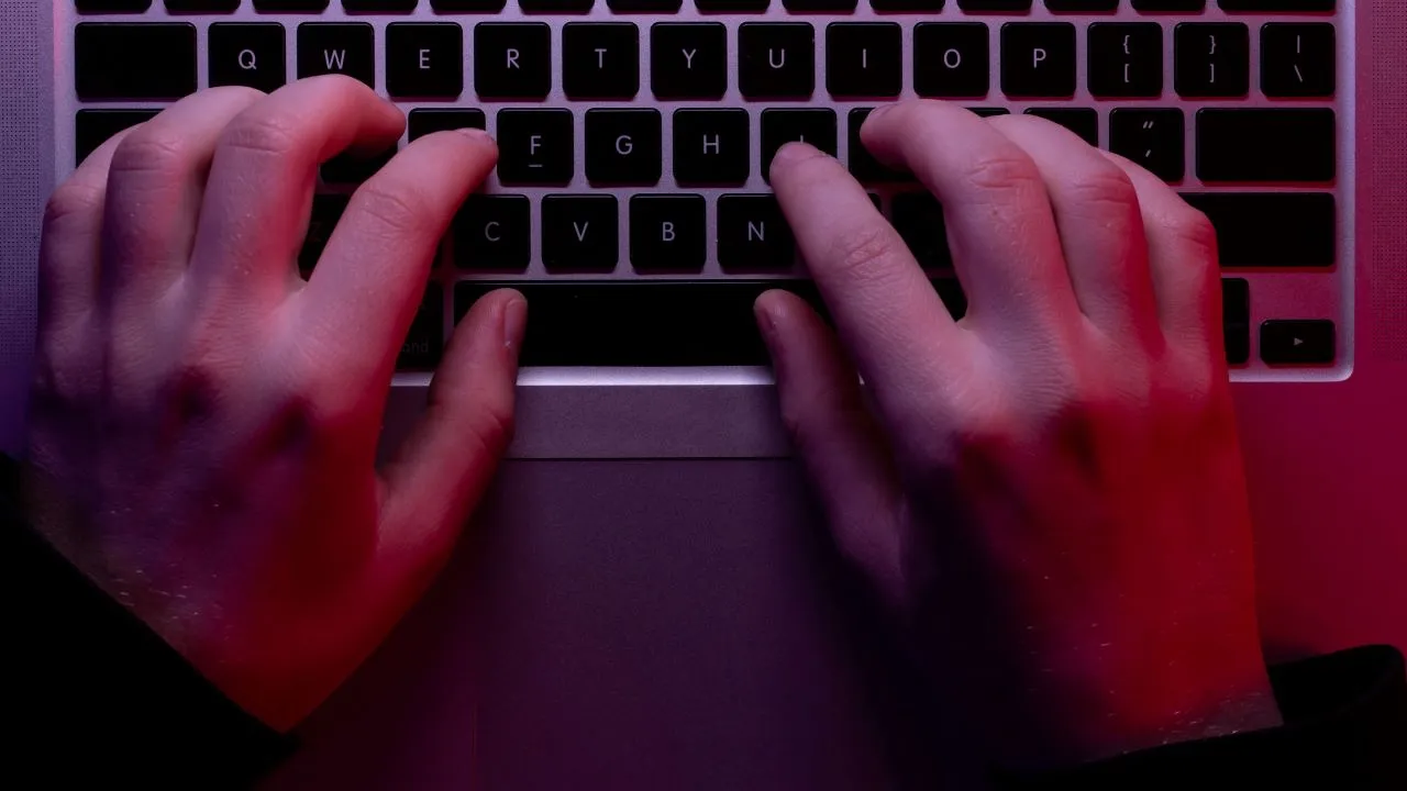 Hands typing on a laptop keyboard under dim red and purple lighting.