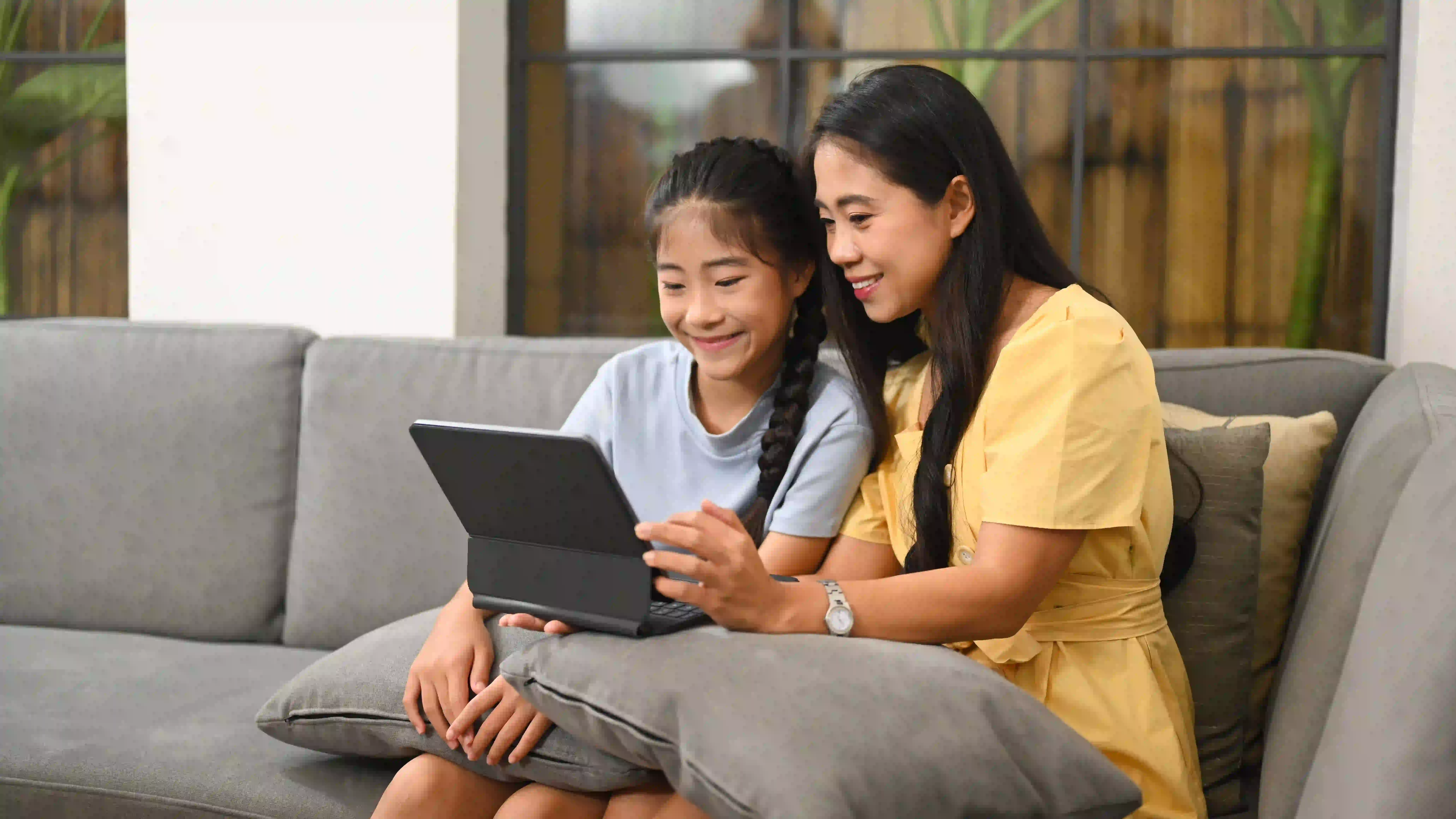 The image shows a mother sitting with her daughter while they are both listening to a teacher teaching on screen.