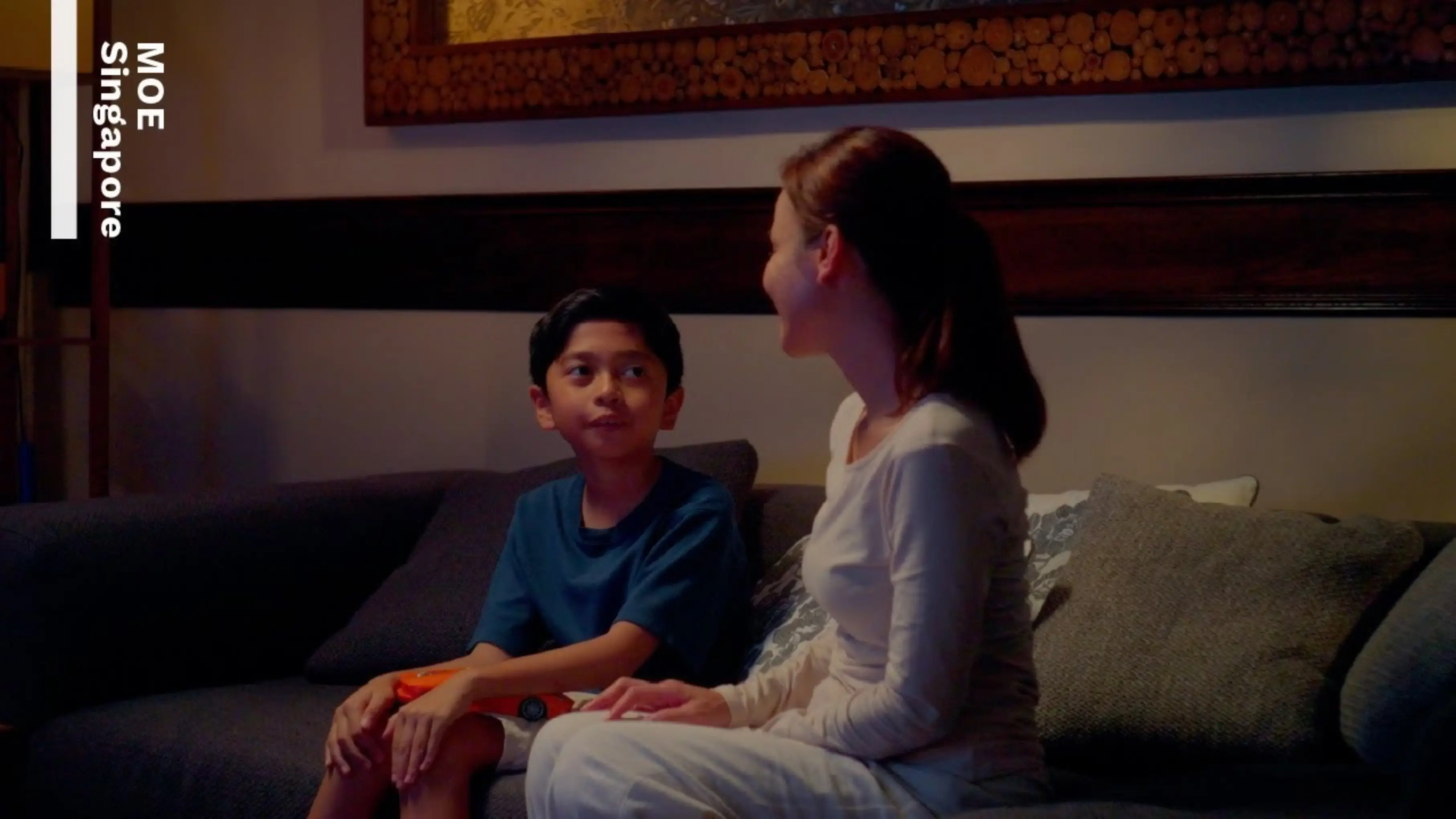 Mother and child sit side by side on a sofa, engaged in a conversation. The mother listening attentively to her child.