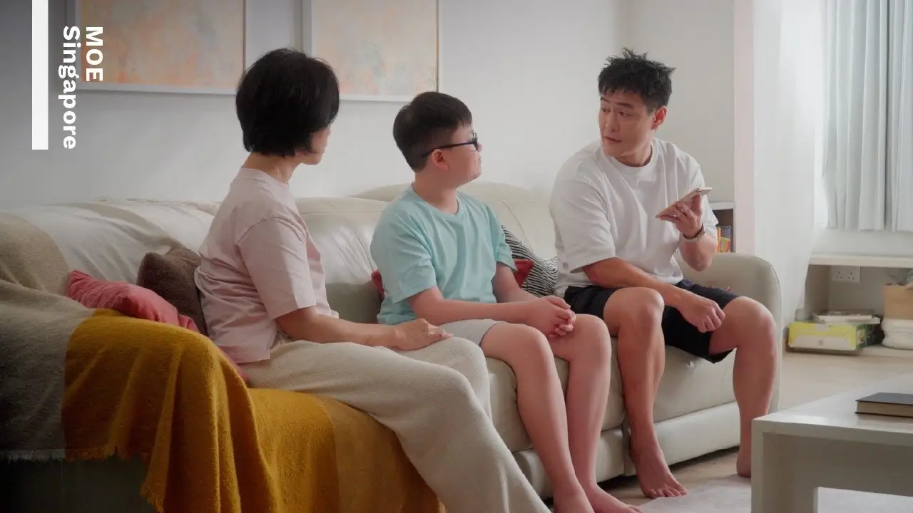 A family sits on a beige sofa. Parents engage in conversation with their young son about getting his first mobile phone.